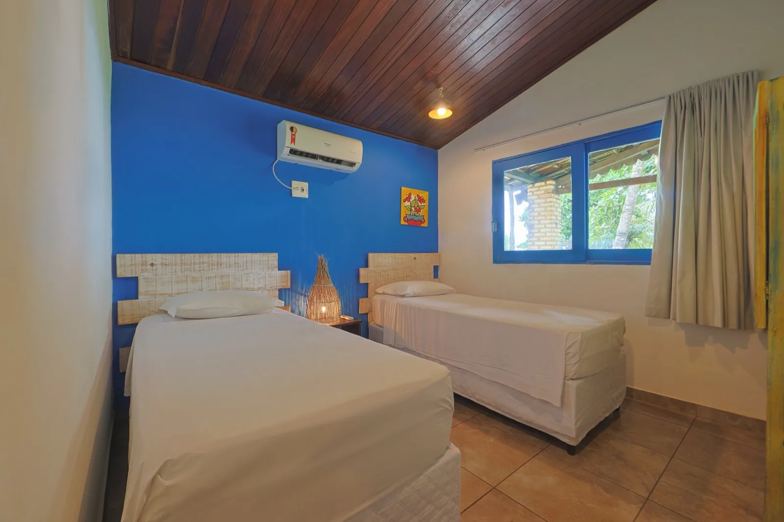 Quarto com duas camas de solteiro, parede azul e ar-condicionado, com janela para a área externa