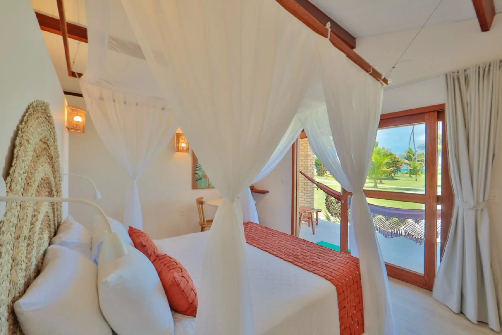 Quarto com dossel branco e vista para a varanda com rede, coqueiral e o mar ao fundo
