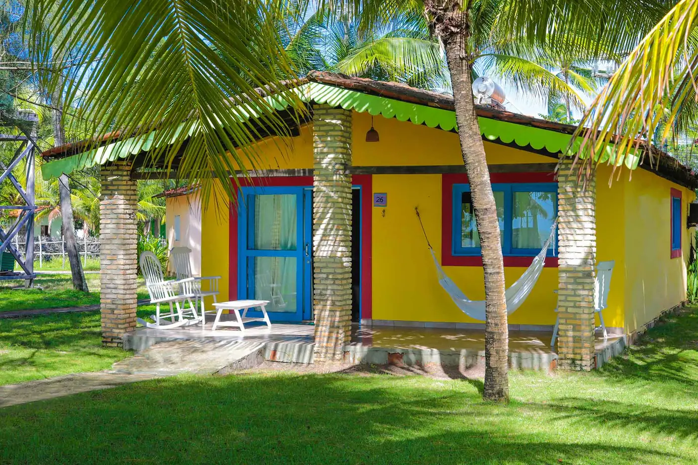Casa amarela com janelas azuis, rede branca e cadeiras de balanço na varanda