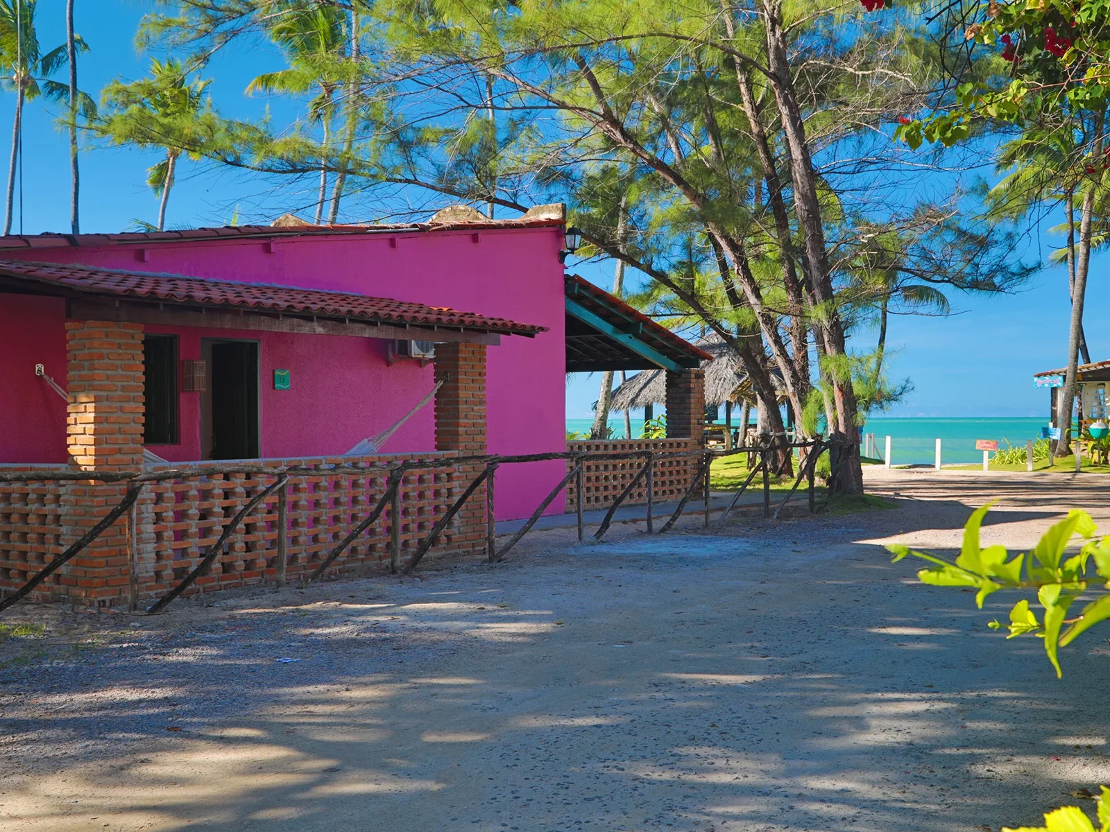 Econômico Sem Vista Mar, chalé rosa com rede na varanda, cerca de tijolo aparente e vista para o mar entre coqueiros