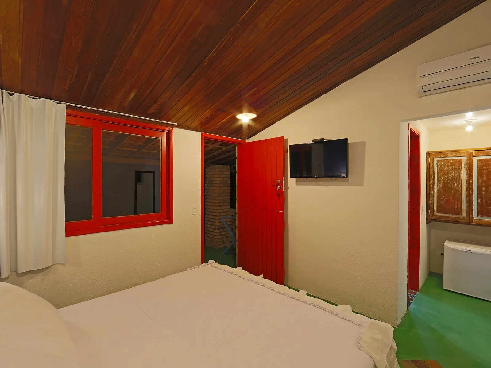 Duplo Econômico Lateral Mar, quarto com cama de casal, porta vermelha, TV de parede e vista para o banheiro com portas rústicas