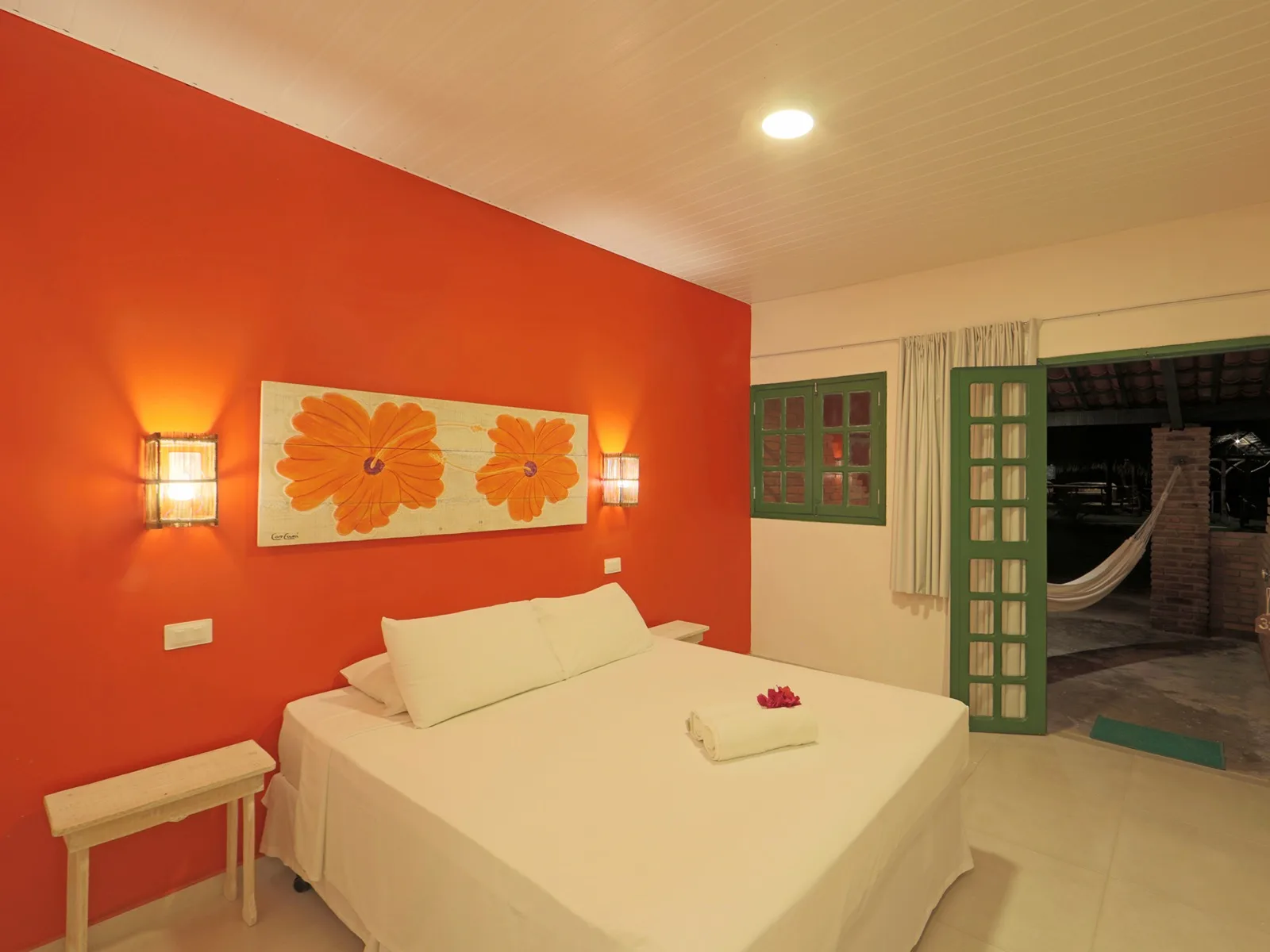 Quarto com parede laranja, painel de flores e cama de casal com toalha e flor sobre o colchão
