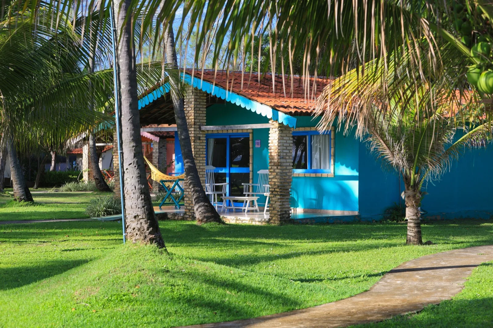 Fachada azul-turquesa da Casa com Vista Mar entre coqueiros, com varanda e cadeiras brancas