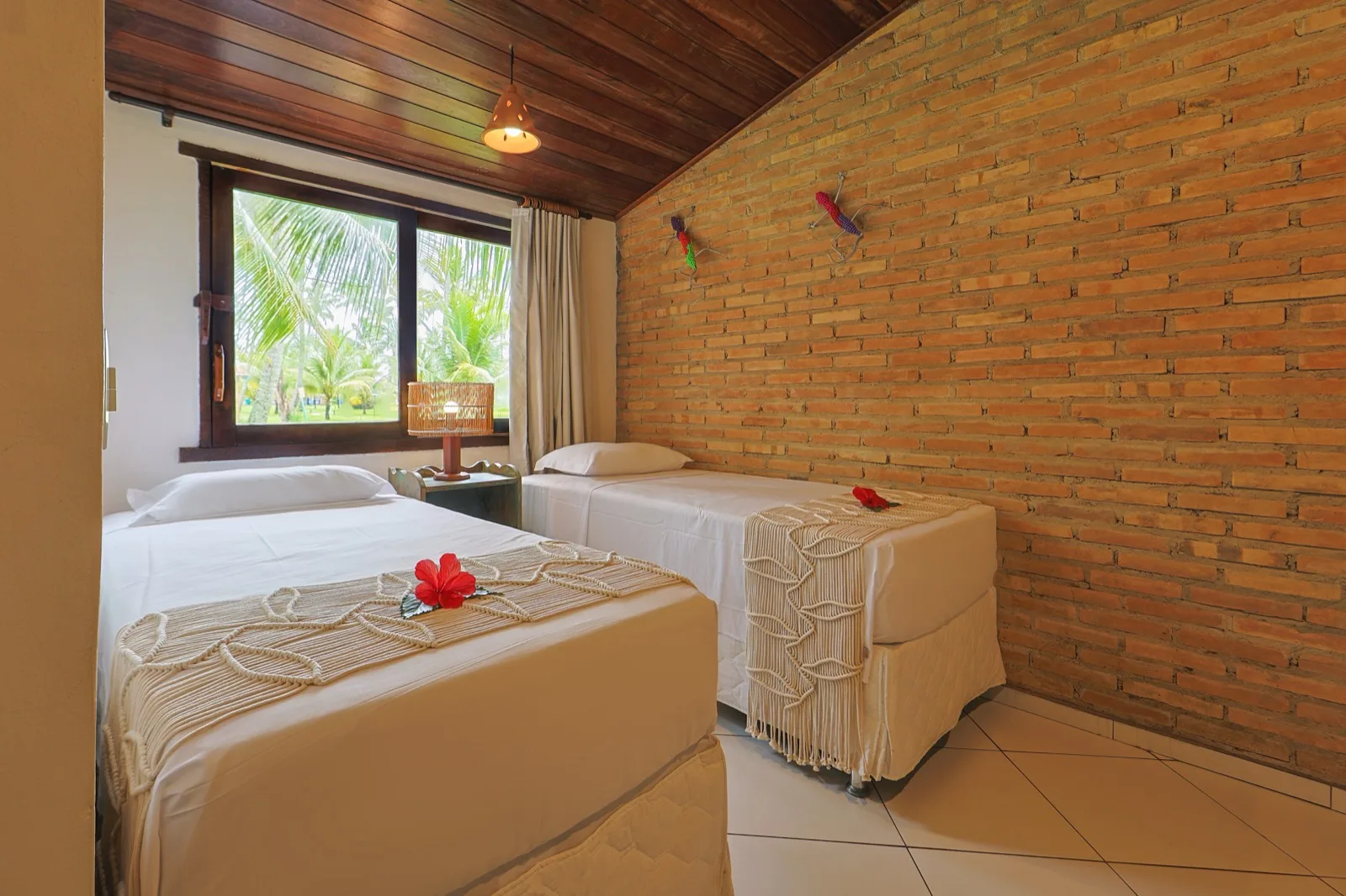 Segundo quarto com cama de casal e cama de solteiro, parede de tijolinhos aparentes e janela com vista para os coqueiros